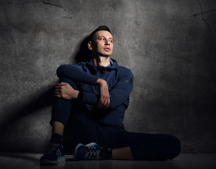 Tired young athletic man in blue sport wear trainsuit sits on the floor by the concrete wall and looks aside