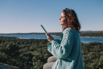 A young brunette woman is reading her tablet in the middle of the nature. She is carefully looking the tablet. She is wearing a blue jacket and black jeans.