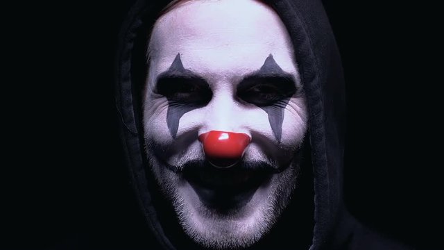 Evil clown smiling to camera against dark background, dangerous maniac in mask