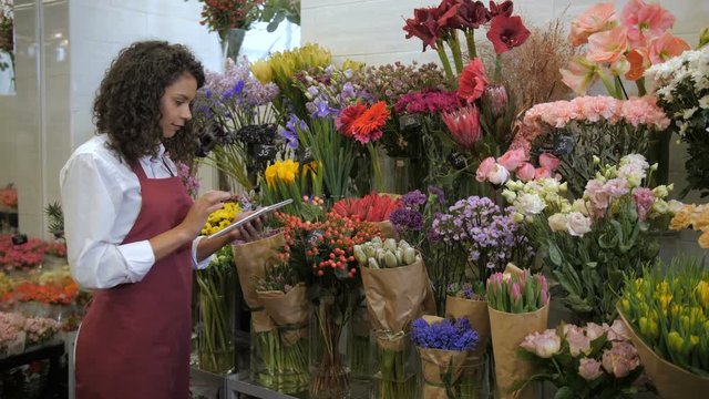 Busy curly mixed race woman florist working on tablet pc in front of stand with fresh cut flowers for sale in floristical shop. Young female checking online quantity and price according to invoice