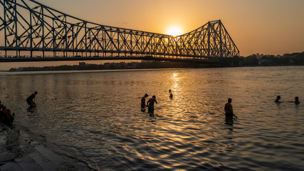 Silhouette of Howrah Bridge at the time of Sunrise.  People bathing and making rituals in the River Ganges.