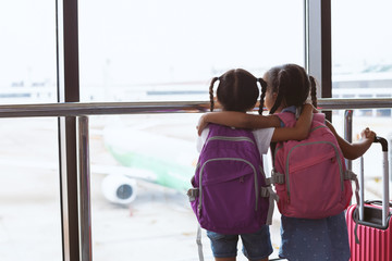 Two cute asian child girls with backpack looking at plane and waiting for boarding in the airport together