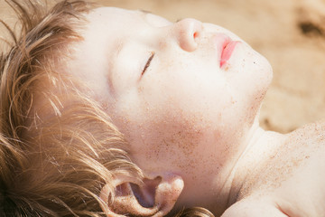 Small child sunbathes on hot sand with his eyes closed in sun.