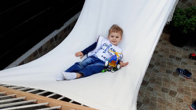 4k video of smiling little boy lying and swinging in hammock. Cild relaxing at house backyard