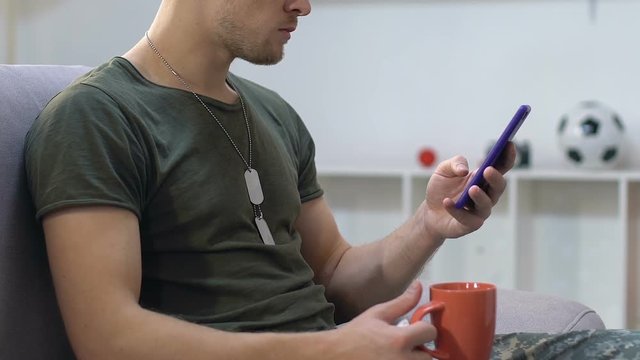Army sergeant reading smartphone messages holding coffee cup, resting on sofa