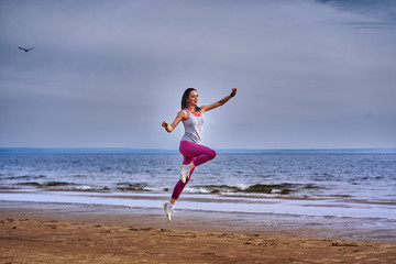 Young woman with black hair jumps while jogging. A woman is engaged in gymnastics in the spring morning on the sandy bank of a large river. Cloudy morning.