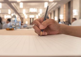 Business agenda briefing shorthand note in seminar workshop or convention hall meeting room with hand holding pencil on notepad for taking note on desk and blur people background - obrazy, fototapety, plakaty