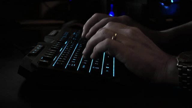 Side view of a cyber criminal's hands typing on a backlit keyboard, dark office