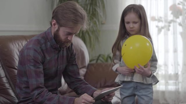 Portrait of concentrated bearded man working with tablet close up. Little girl unsuccessfully trying to give the man the balloon but irritated father shows her that he is busy. Child is sad