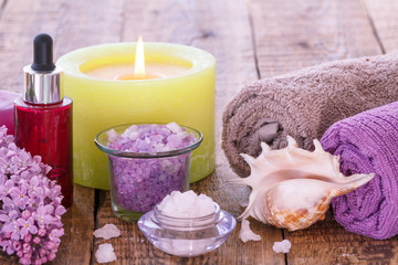 Fototapeta na wymiar Burning candle, bowls with sea salt, bottle with aromatic oil, lilac flowers and towels on wooden background.