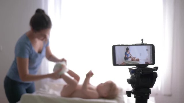 educational video blog for young moms, young mom vlogger changes diaper to small child boy while recording education video for followers live on smartphone at home