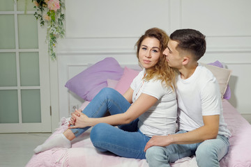 Young couple in love man and woman in white t-shirts and jeans are sitting on bed in the bedroom, man is looking at woman, but she is looking at camera all time.