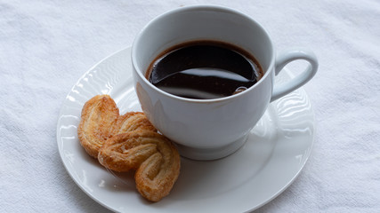 Cup of Greek or Turkish coffee, on small white saucer plate, with two cookie pastries, on white cloth surface