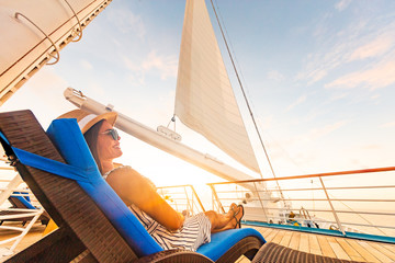 Luxury cruise vacation woman relaxing in lounger chair enjoying sunset on yacht deck with sail in wind sailing in getaway destination summer travel lifestyle.