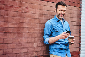 Cheerful man leaning against wall in the city using smartphone and having coffee