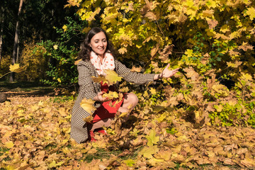 Young brunette woman wearing red dress throwing autumn yellow maple leaves up. Happy girl enjoying weekend outdoor walking. Sunny day and fall nature on background.