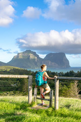 Fototapeta na wymiar Evening sunset view of subtropical Lord Howe Island in the Tasman Sea, belonging to Australia. Mt Lidgbird and Mt Gower in background.Rear view of beautiful young female hiker with backpack at a fence