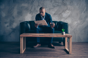 Portrait of his he nice stylish classy chic imposing elegant attractive guy professional top manager company banker sitting on couch reading paper at workplace station gray concrete wall