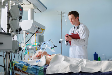 Intensive care caucasian doctor examines intubated critical stance patient writing notes to case...