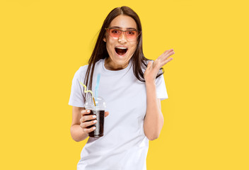 Beautiful female half-length portrait isolated on yellow studio background. Young smiling woman in red sunglasses with drink. Facial expression, summer, weekend, resort concept. Trendy colors.