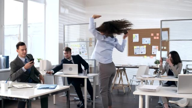 Beautiful young businesswoman holding smartphone and dancing in the center of office while colleagues smiling and taking pictures of her with phones
