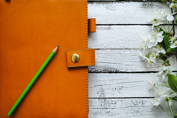Orange notebook and green pencil lie on a white wooden background between the branches with the flowers of cherry