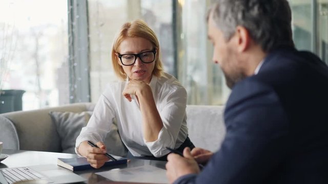 Business partners cheerful man and woman are talking in cafe looking at papers sitting at table with laptop. Professional conversation and people concept.