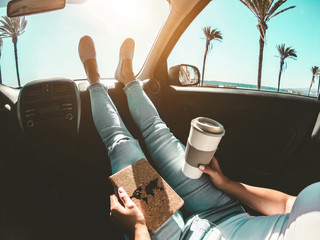 Woman drinking coffee paper cup inside car with feet on dashboard - Girl relaxing in auto trip reading travel book with ocean beach and palms in background - Traveler concept - Focus on hands