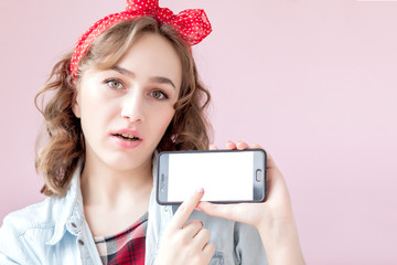 Naklejka premium Beautiful young woman with pin-up make-up and hairstyle over pink background with mobile phone with copy space