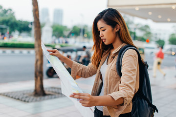 Pretty casual Asian woman with backpack reading paper map while exploring city of Bangkok, Thailand