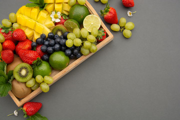 Various fresh fruits - mango, grapes, tangerine, lime, strawberry, kiwi, mint, on wooden tray and a gray background. Copy space.