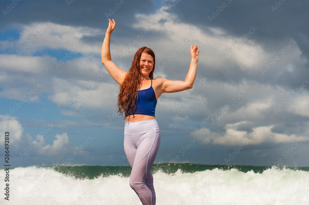 Wall mural young happy and attractive red hair woman having fun standing on rock at the sea enjoying beach holi