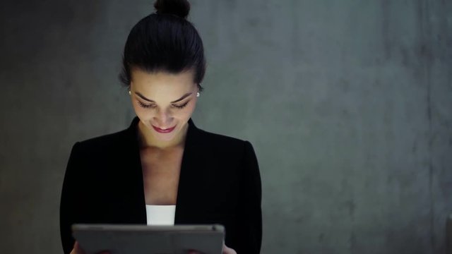 Young business woman with tablet walking in office, concrete wall in background.