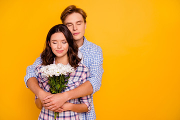 Close up photo of pair in love with overjoyed faces he him his she her lady boy flowers in hands holding each other in hugs eyes closed wearing casual plaid shirt outfit isolated on yellow background