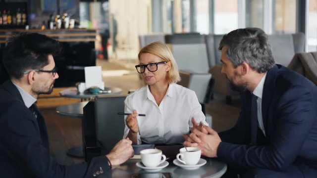 Cheerful business lady with blond hair is negotiating agreement with male partners bearded men during meeting in cafe. Talks, communication and work concept.