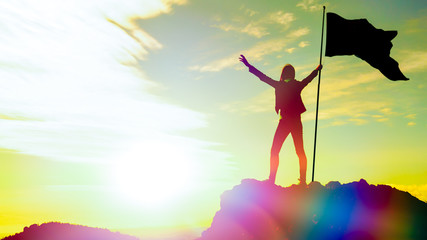 High achievement, silhouettes of the girl, flag of victory on the top of the mountain, hands up. A man on top of a mountain. Conceptual design. Against the dramatic sky with clouds at sunset.