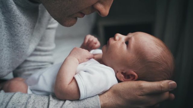 Dad in grey sweater kisses his newborn baby on the nose. Tiny baby in white outfit rests in the arms of its father staring up at him. Love between parent and child. Slow motion.