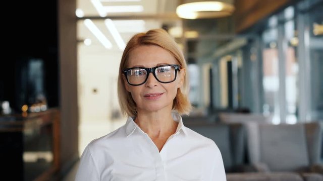 Portrait of beautiful senior businesswoman with blond hair wearing glasses looking at camera standing in cafe alone. Business people and leadership concept.