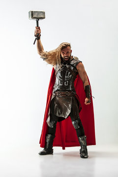 Long hair and muscular male model in leather viking's costume with the big hammer cosplaying Thor isolated on white studio background. Full-lenght portrait. Fantasy warrior, antique battle concept.