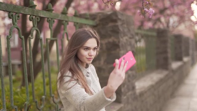 Young Caucasian Female in Gary Jacket Standing By Fence of Park on Street and Taking Selfies With Smartphone. Light Camera Movement