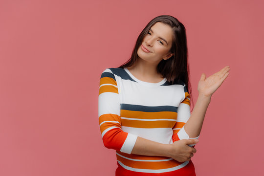Lovely woman demonstrates something with palm on free space, wears striped casual jumper, tilts head, has pleased expression, models over pink background, suggests going in this direction, shows way