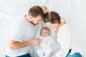 overhead view of happy parents lying on white sheet and looking at baby