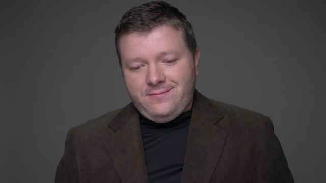 Portrait of shy brunette middle-aged businessman in suit smiles into camera being embarrassed on gray background.