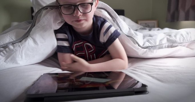 A six years old boy reading a computer tablet while lying in his bed.