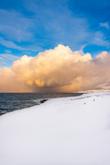 Snow and sea: the snow arrives all the way to the shoreline of an unfrozen deep blue sea while the clouds in the blue sky turn yellow as the sunset approaches
