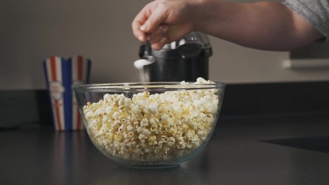 Seasoning a freshly popped bowl of warm and tasty popcorn. Woman making popcorn in his kitchen. Family movie time evening snack.