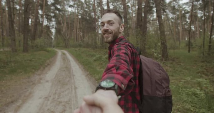 Happy bearded man dressed in red checked shirt holding POV hand and walking in ht forest