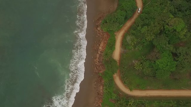 Aerial view of car driving on interesting dirt road between ocean waves and central american jungle. car drives top of scene to bottom and exits right