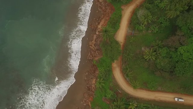 Aerial view of car driving on interesting dirt road between ocean waves and central american jungle. Car approaches scene from right.