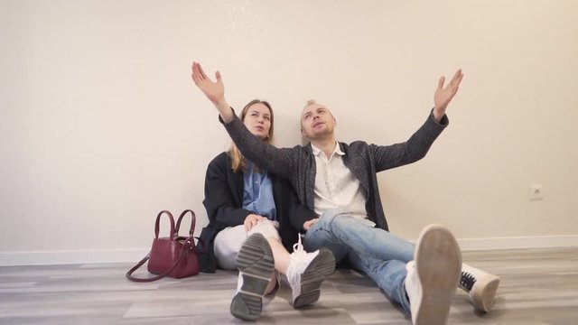Young Happy Caucasian Married Couple Sitting on Floor of New Empty Aprartment Real Estate Property and They Talking About Arragement and Future Look of Room. Light Camera Movement